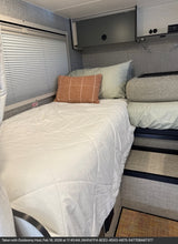 Cozy RV rental interior featuring a comfortable bed with white bedding, pillows, window blinds, and overhead storage.