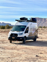 White Ford Transit RV rental with custom graphics & off-road gear, parked in a desert mountain landscape.
