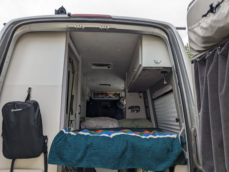 RV rental: Step inside this cozy camper van. Rear view showing a comfortable bed, storage, and shower for travel.
