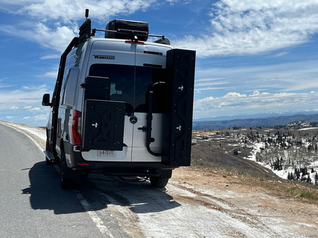 A white Winnebago rv rental van with overland gear is parked on a mountain road, overlooking snowy valleys and distant peaks.