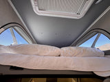 Inside this RV rental: a comfy bed with white pillows & blanket. Overhead skylight, screened windows reveal blue sky & hills.