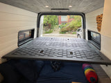 Spacious RV rental van interior with a modular bed platform over cozy bedding, light paneling, and a wood ceiling.