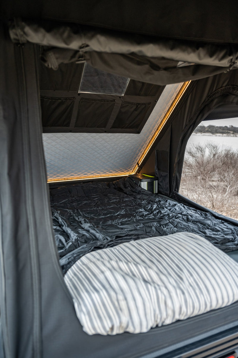 Inside the rooftop tent looking out the open door, showing the upgraded mattress with bedding, the quilted insulated ceiling panel, and the warm interior LED light strip running along the roofline — photo 4