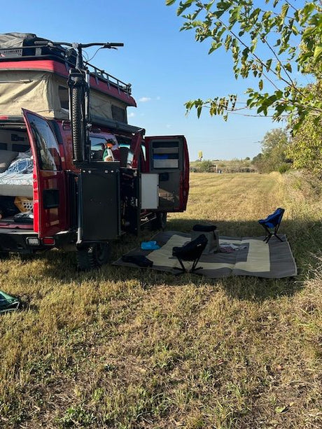 This red RV rental camper van with open doors, bike, and pop-top is set up with an outdoor mat and chairs in a grassy field.