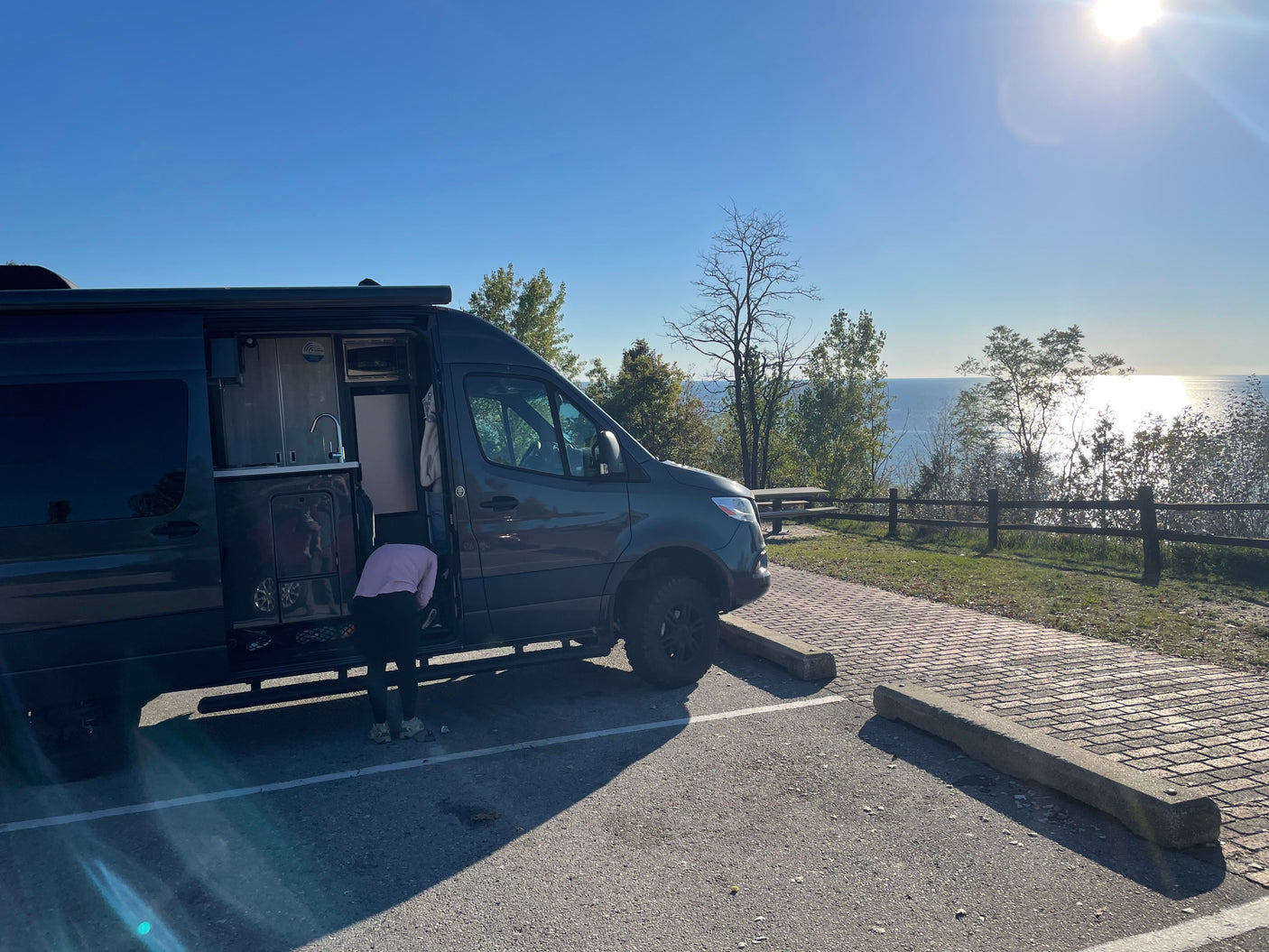 A dark gray rv rental camper van, with its side door open to a kitchenette, is parked overlooking a bright, sunny ocean.
