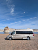 RV rental: A tan Roadtrek Zion camper van parked by a scenic lake and impressive rock formations under a clear blue sky.