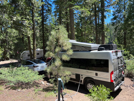 RV rental & Tesla EV parked at a sunny forest campsite with a travel trailer. Connected to utilities, ready for adventure.