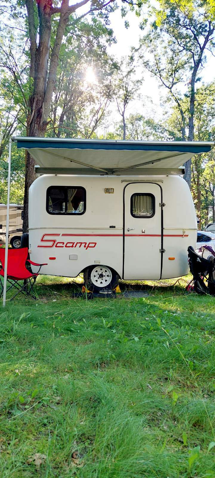 A Scamp travel trailer RV rental, with awning extended, is parked on grass in a sunny campground, ready for a camping adventure.