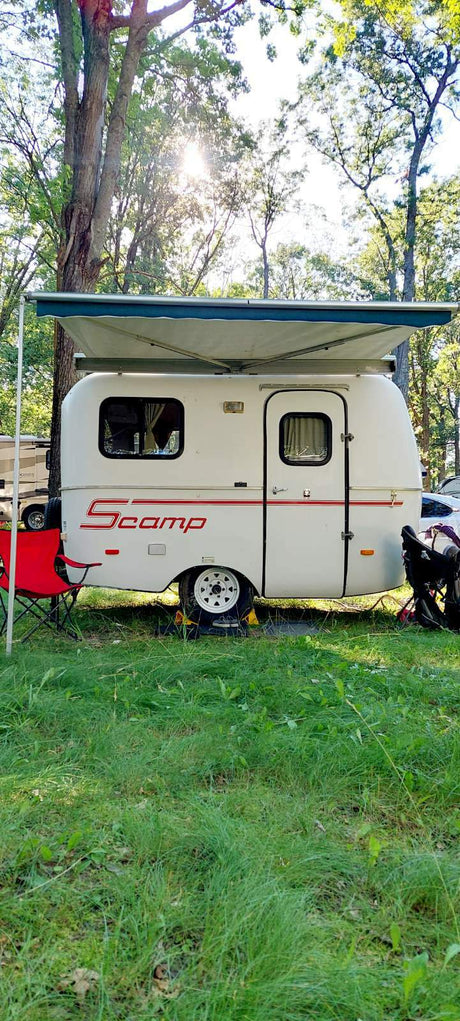 A Scamp travel trailer RV rental, with awning extended, is parked on grass in a sunny campground, ready for a camping adventure.