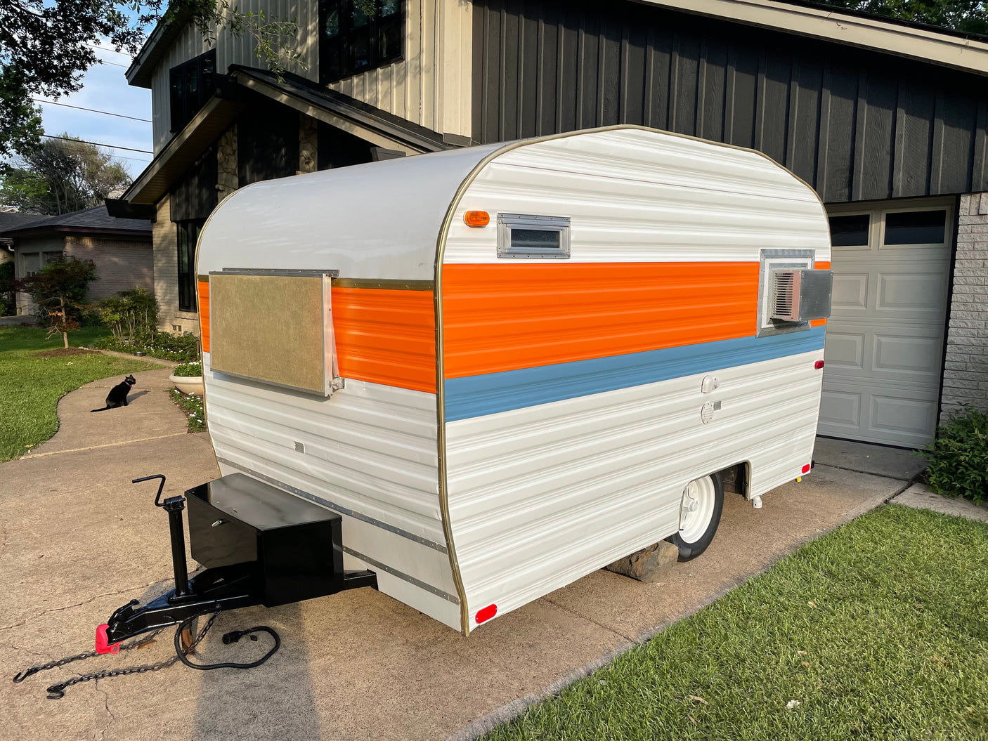 A colorful, retro-style rv rental camper, white with orange and blue stripes, sits on a driveway. A black cat is nearby.