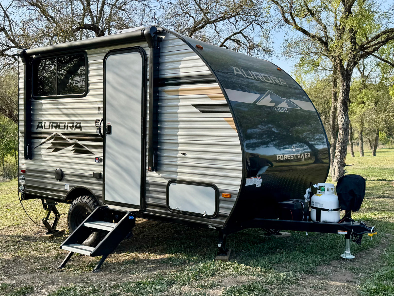 A sleek Forest River Aurora travel trailer, ideal for your next rv rental, parked on grass amidst mature trees.