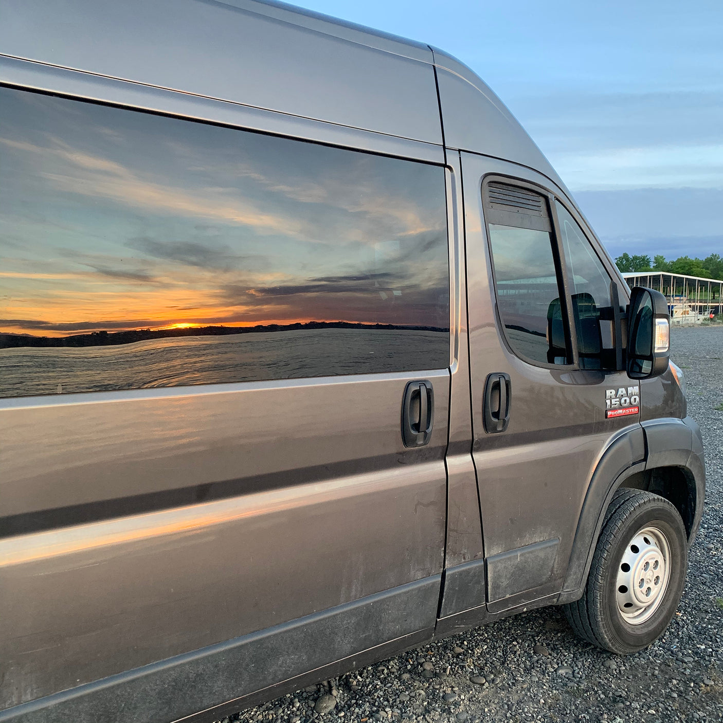 This grey Ram ProMaster for rv rental reflects a stunning sunset over water. Perfect for van life adventures, day or night.