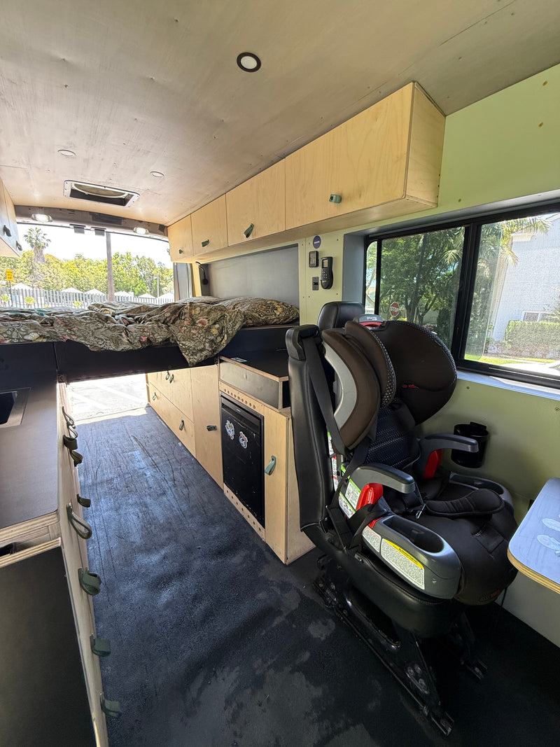 Inside this RV rental: a cozy bed, child car seat, mini-fridge, and custom cabinetry for family travel comfort. — photo 11
