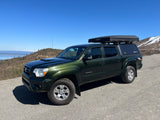 This green Toyota Tacoma, equipped with an iKamper tent & SmartCap, offers a unique adventure, akin to an RV rental in the mountains.