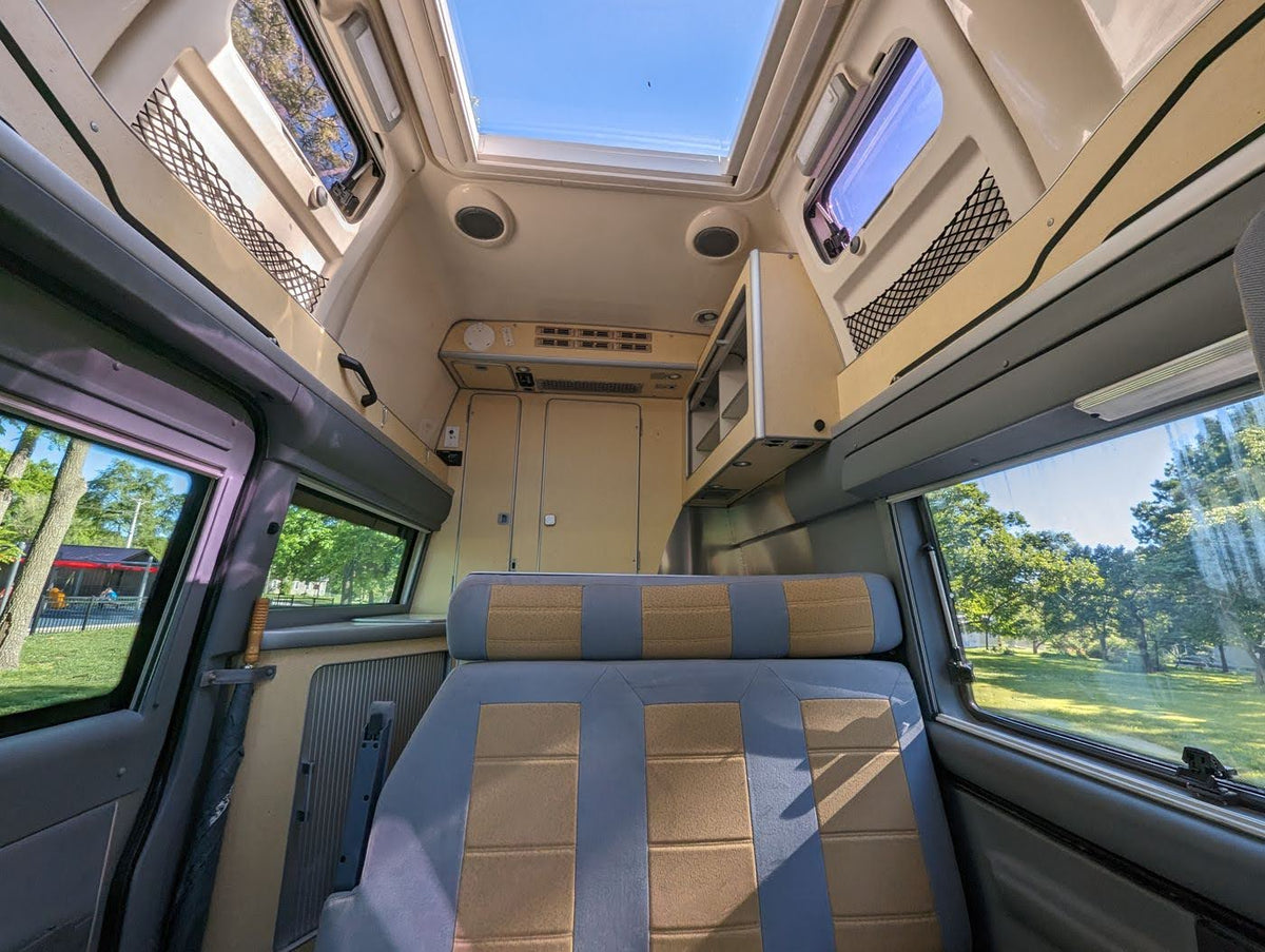 Bright RV rental camper van interior: spacious seating, skylight, and large windows reveal a sunny, treed outdoor view. — photo 7