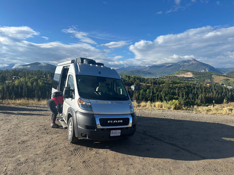 An RV rental camper van stopped on a mountain overlook. A person accesses the van, revealing a breathtaking view of forested peaks and blue sky.