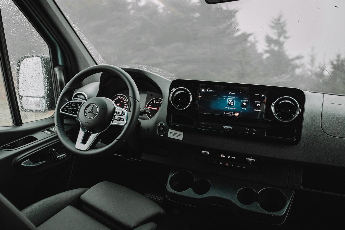 Interior of a Mercedes rv rental: dashboard, steering wheel, and infotainment system. Rain spatters the windshield, showing blurred trees.