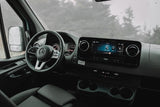 Interior of a Mercedes rv rental: dashboard, steering wheel, and infotainment system. Rain spatters the windshield, showing blurred trees.