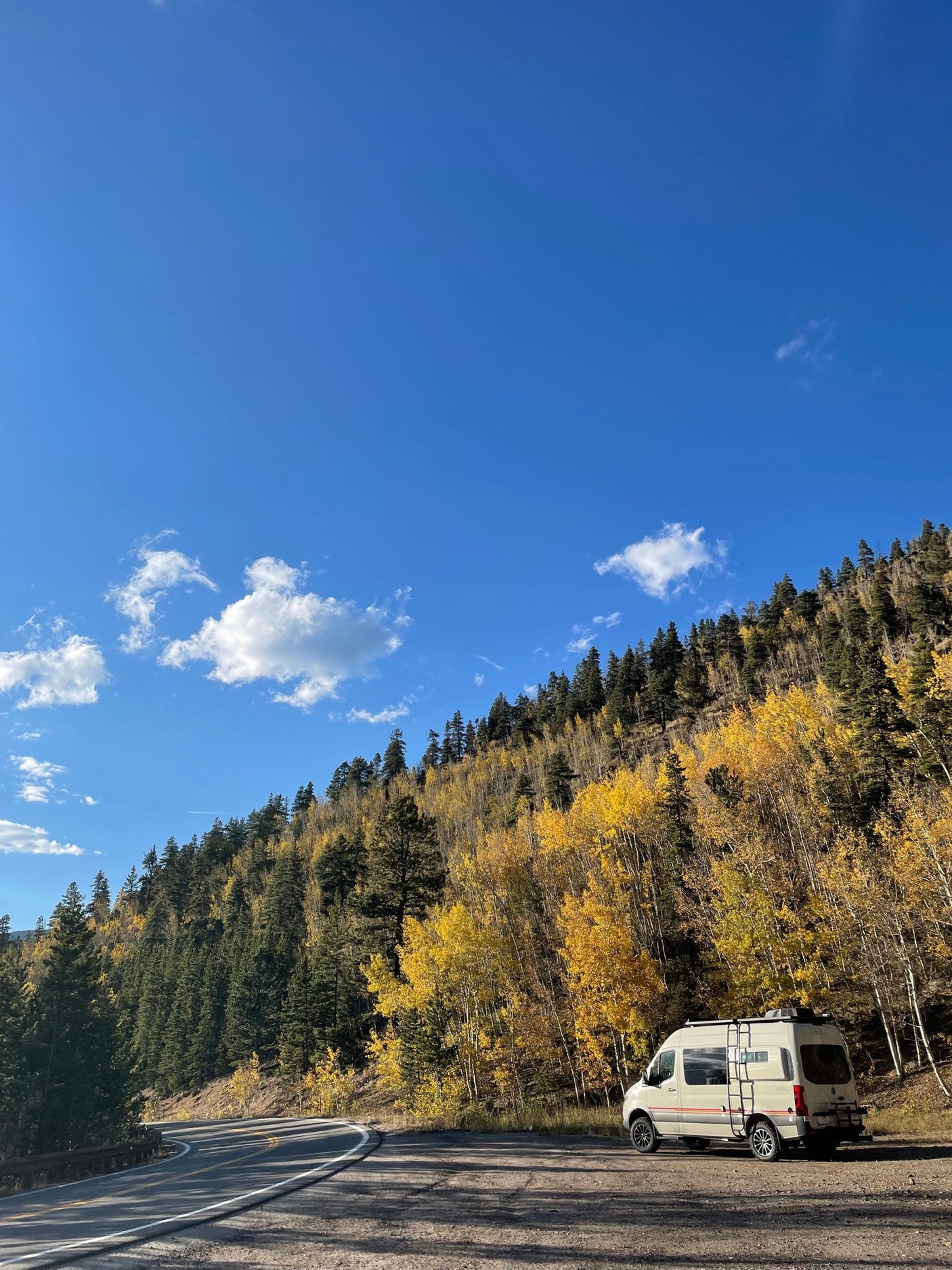 An RV rental is parked on a scenic mountain road surrounded by vibrant yellow and green autumn trees under a clear blue sky.