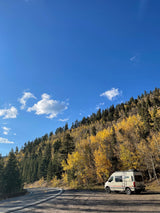 An RV rental is parked on a scenic mountain road surrounded by vibrant yellow and green autumn trees under a clear blue sky.