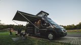 This RV rental camper van is set up for a relaxing campsite adventure, with an awning extended & pop-top raised.