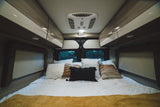 Cozy RV rental bedroom featuring comfortable bedding, overhead cabinets, a ceiling AC unit, and rear window view.