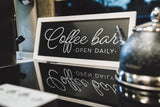 Charming RV rental coffee bar setup: a black 'Coffee bar - OPEN DAILY -' sign with reflection, alongside a kettle and appliance.