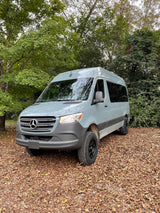 A light grey Mercedes-Benz Sprinter van, ideal for rv rental, is parked on brown autumn leaves amidst green trees.