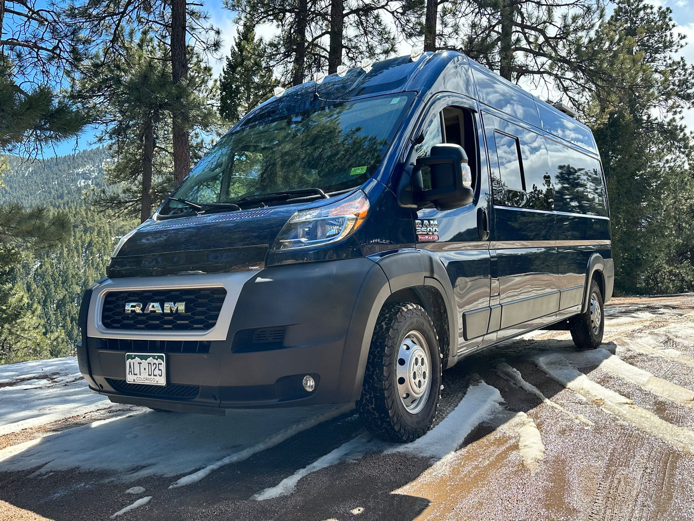 Dark blue Ram ProMaster campervan, perfect for an RV rental adventure, parked on a snowy forest road with mountains.