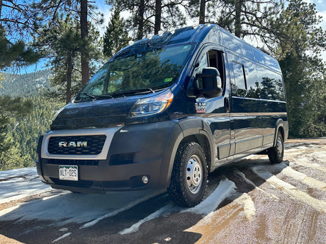 Dark blue Ram ProMaster campervan, perfect for an RV rental adventure, parked on a snowy forest road with mountains.