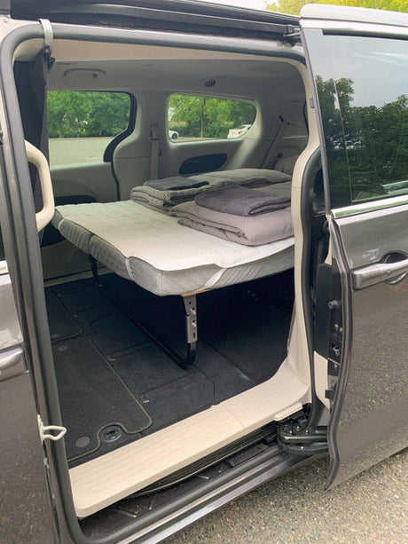 A gray minivan's open door shows an elevated bed with blankets, perfect for an RV rental or van camping trip.