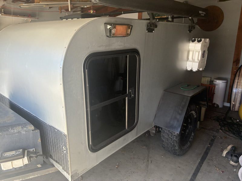 Street-side profile of the teardrop showing the sliding-window entry door, roof-mounted marine light, twin white water jugs and the diamond-plate lower skirt — photo 2
