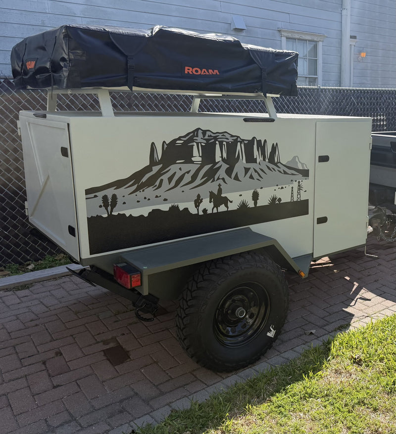 Homemade 2025 overland trailer in cream and sage green, rear driver-side three-quarter view, with a black Roam Vagabond rooftop tent closed on the roof rack, a desert mountain and horseback-rider vinyl decal down the side panel, lockable hatch doors and an off-road tire on a black steel wheel — photo 1