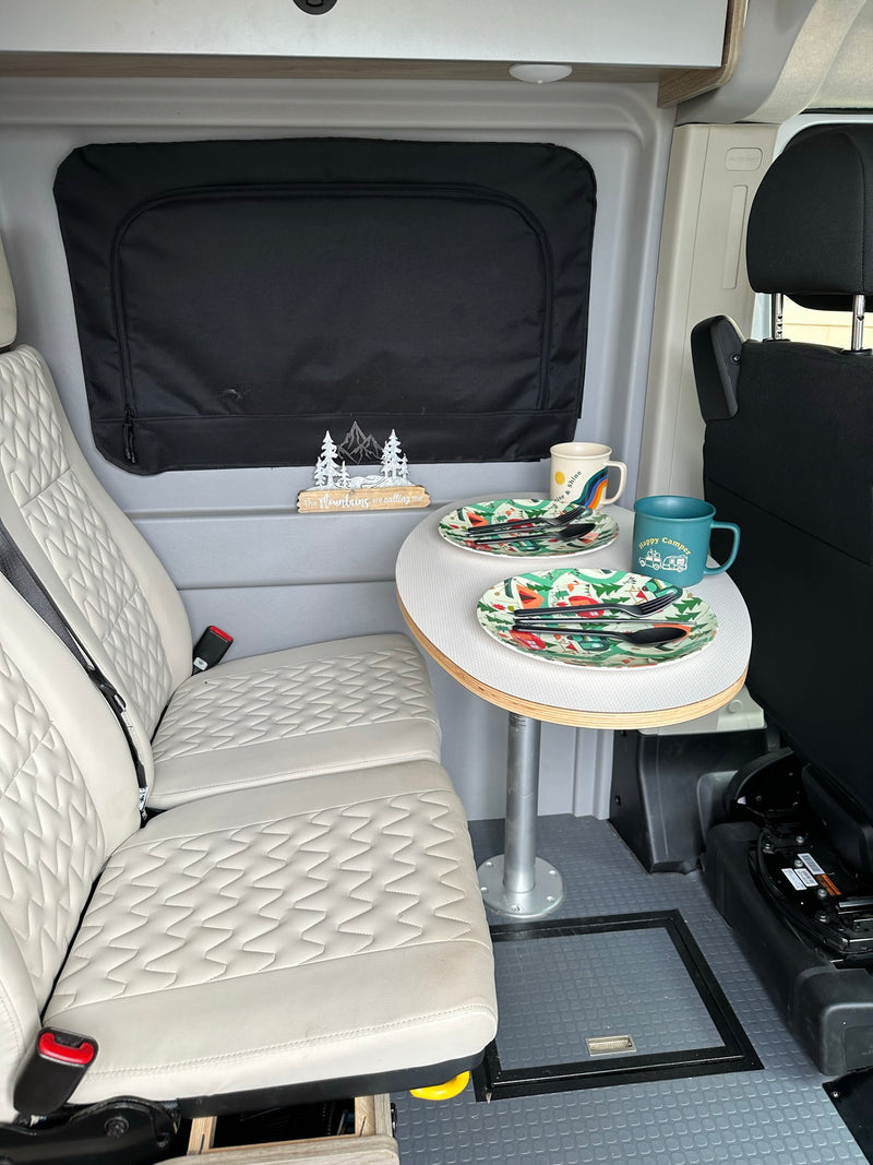 RV rental dining space: white quilted seats, round table with camping plates/mugs, and mountain decor under a black window shade. — photo 19