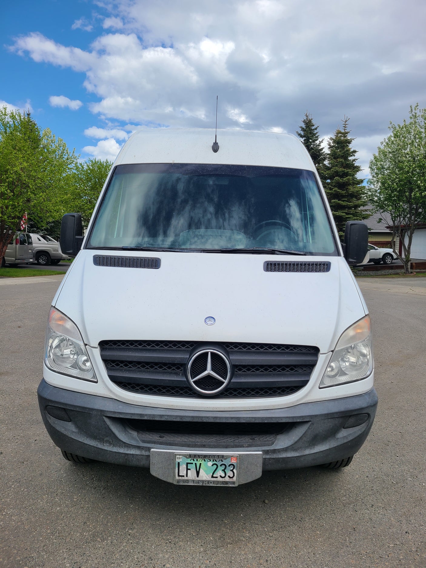 Front view of a white Mercedes-Benz Sprinter van, a potential RV rental, with an Alaska license plate. Outdoor scene.