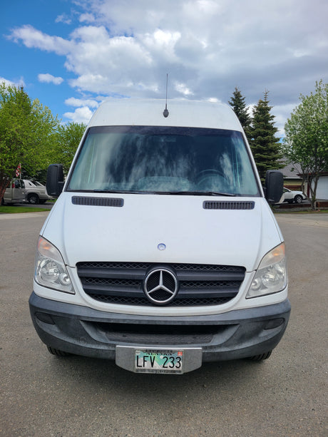 Front view of a white Mercedes-Benz Sprinter van, a potential RV rental, with an Alaska license plate. Outdoor scene.