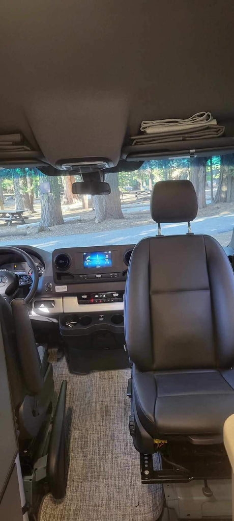 RV rental interior: Front cabin with dashboard, swiveling passenger seat, and view of a treed campground.