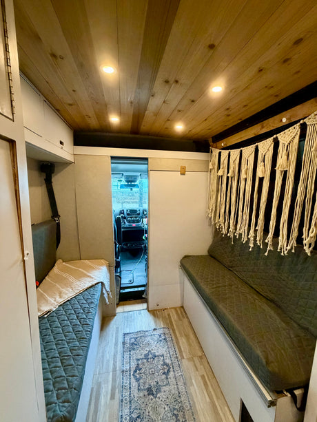 Cozy RV rental camper van interior. Features a bright wood plank ceiling with lights, cushioned benches, and a boho macrame wall hanging.