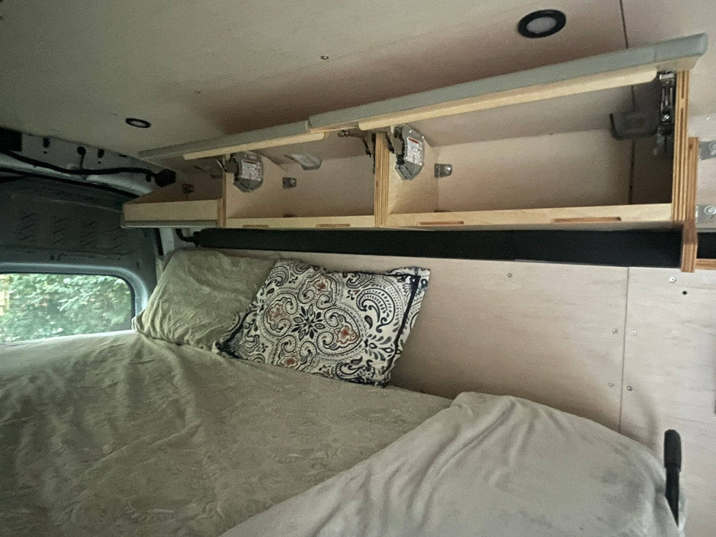 RV rental interior featuring a bed with pillows, open overhead storage, and a window view of trees. — photo 6