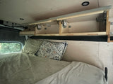 RV rental interior featuring a bed with pillows, open overhead storage, and a window view of trees.