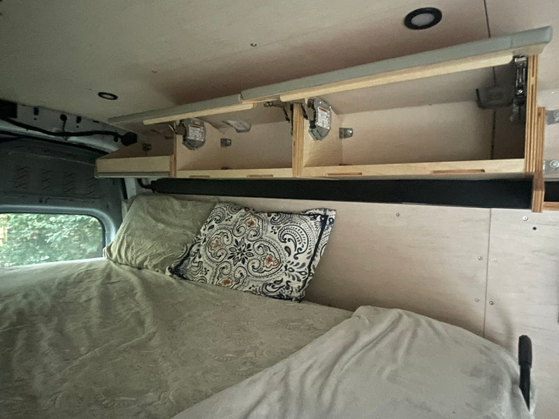 RV rental interior featuring a bed with pillows, open overhead storage, and a window view of trees. — photo 6