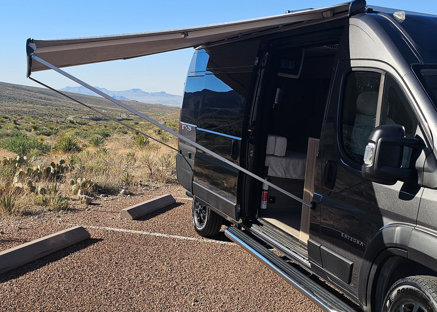 A dark grey camper van with an extended awning is parked in a desert landscape, ideal for an rv rental adventure.