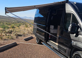 A dark grey camper van with an extended awning is parked in a desert landscape, ideal for an rv rental adventure.