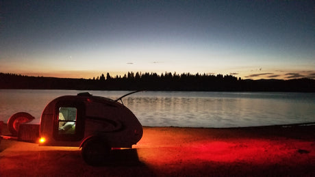 An RV rental teardrop camper glows by a tranquil lake at sunset, trees silhouetted against the colorful sky.