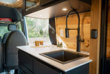 Inside a stylish RV rental, featuring a sleek kitchen counter with a black sink & faucet, wooden walls, and soft lighting.