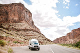 White Ford RV rental van on a scenic desert road, framed by towering red rock canyons under a cloudy sky.