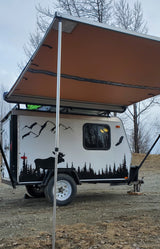Customized small camper trailer with an extended awning, featuring nature decals of moose, trees, and birds. Ideal for an RV rental or camping trip.