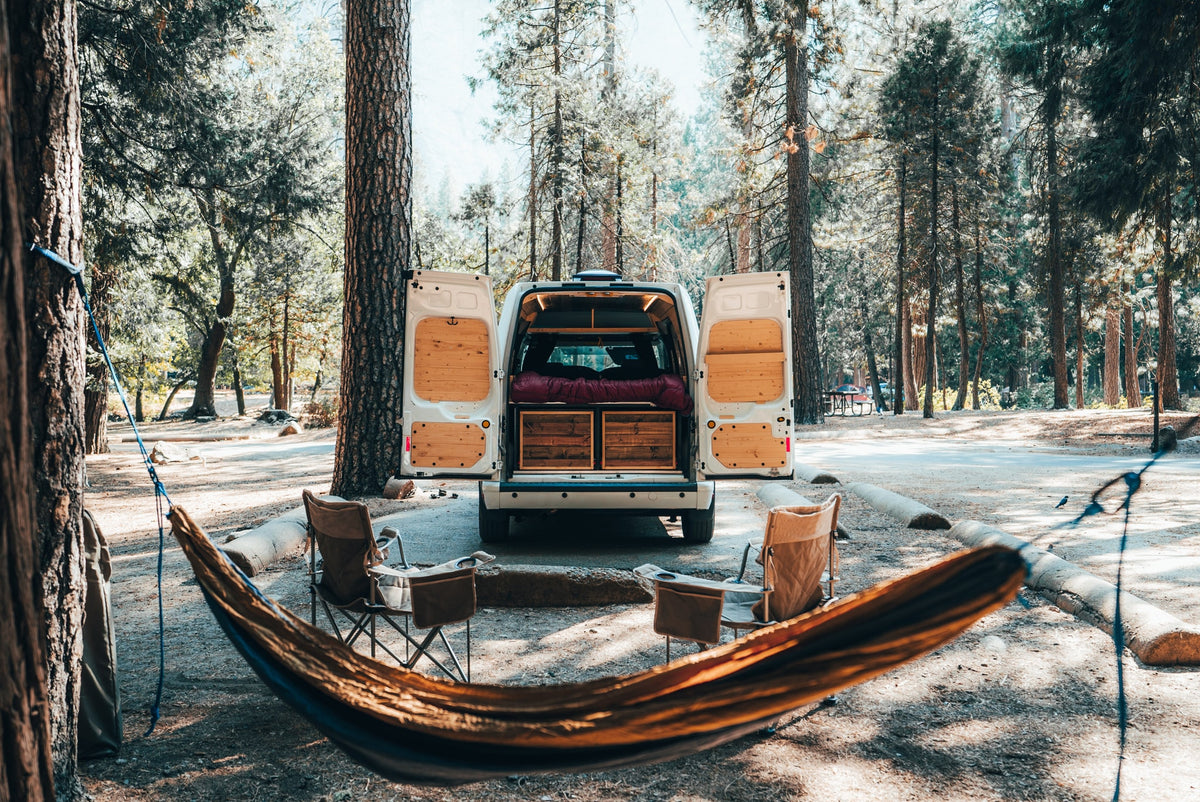 Experience the outdoors! This RV rental camper van setup in a forest campsite features a hammock & chairs for relaxation.|RV rental trip: Man cooks from his camper van's pull-out kitchen