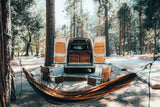 Experience the outdoors! This RV rental camper van setup in a forest campsite features a hammock & chairs for relaxation.