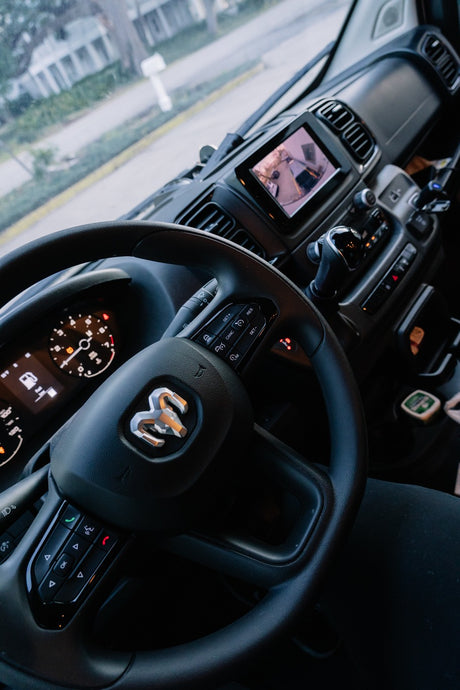 Inside a modern RV rental van: steering wheel with Ram logo, dashboard, and infotainment screen displaying a camera view.